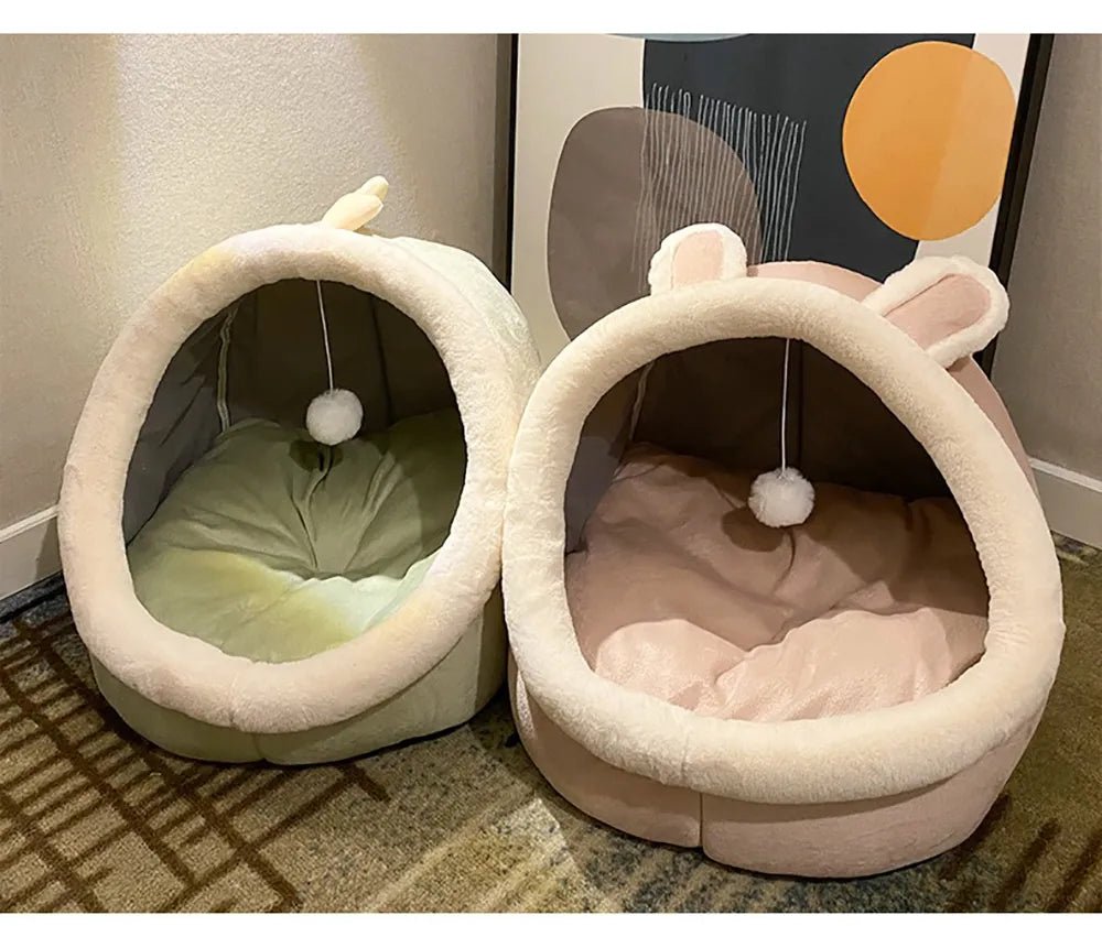 Plush cat beds with hoods and pompom toys, one green and one pink, in a modern room