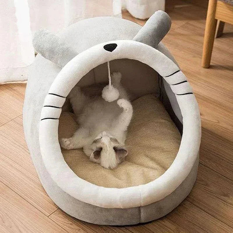 White cat lying inside a plush animal-shaped cat bed with hanging toy on wooden floor