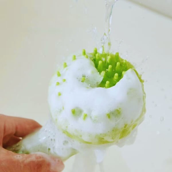Green silicone pet bath brush with soft bristles and soap suds under running water