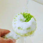 Green silicone pet bath brush with soft bristles and soap suds under running water