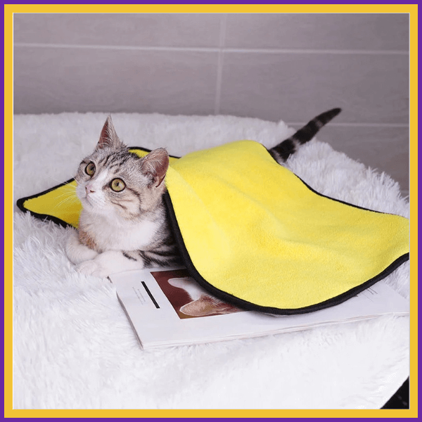 Cat lying under a yellow microfiber towel on a fluffy white surface